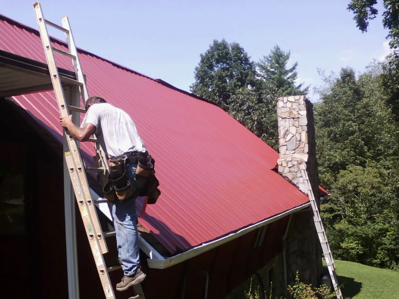 Roofer on a ladder beside a red metal roof and stone chimney — Soffit & Fascia Repair by Roofing Brothers in Briceville